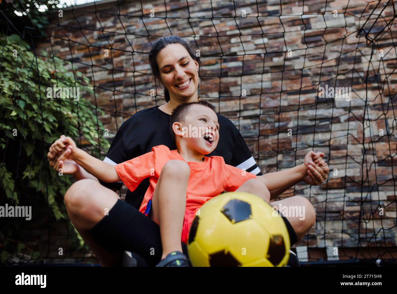 Mom playing football with her son, dressed in football jerseys. The family as one soccer team. Family sports activities outside in the backyard or on Stock Photo