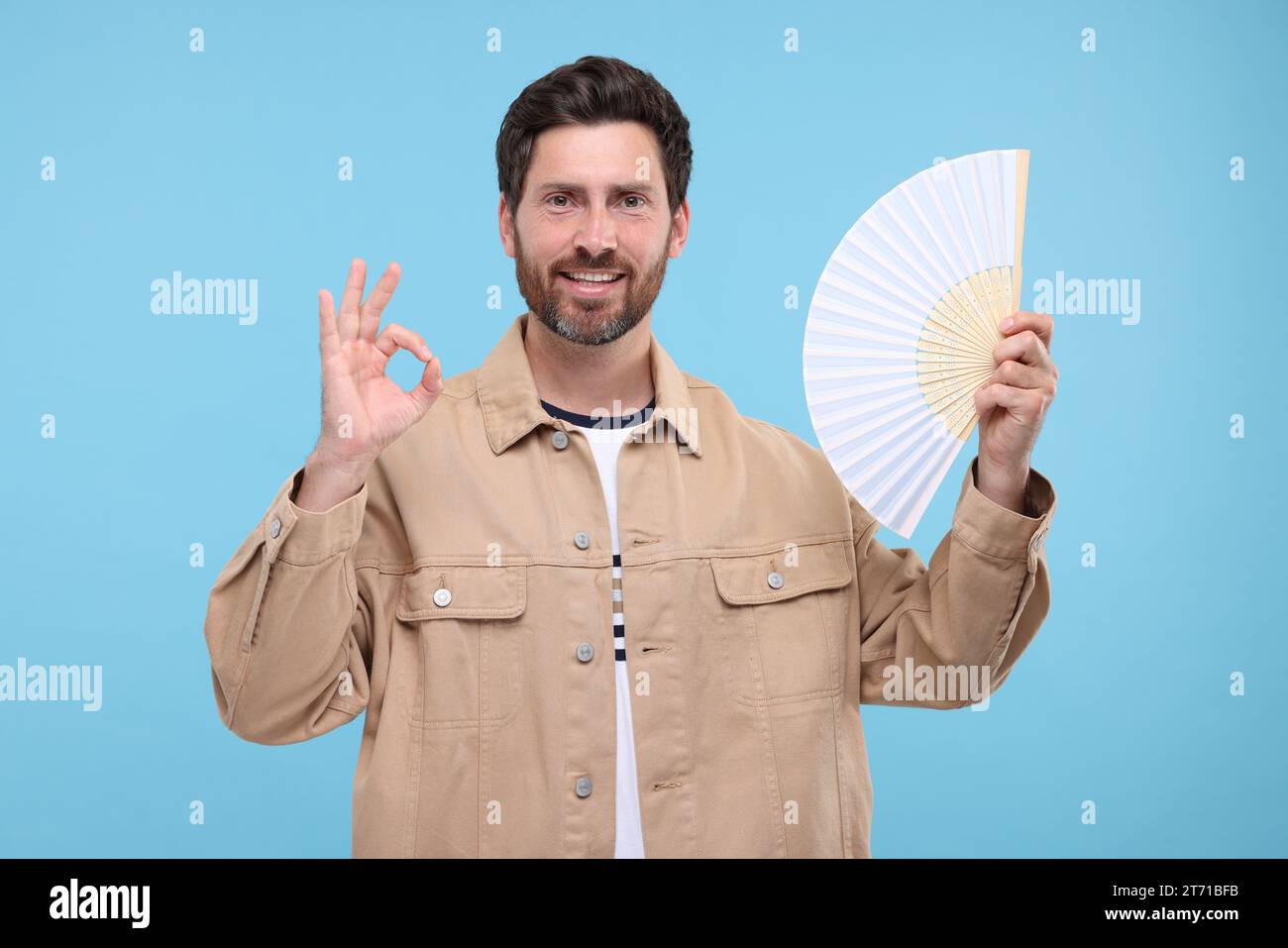 Happy man holding hand fan and showing ok gesture on light blue ...