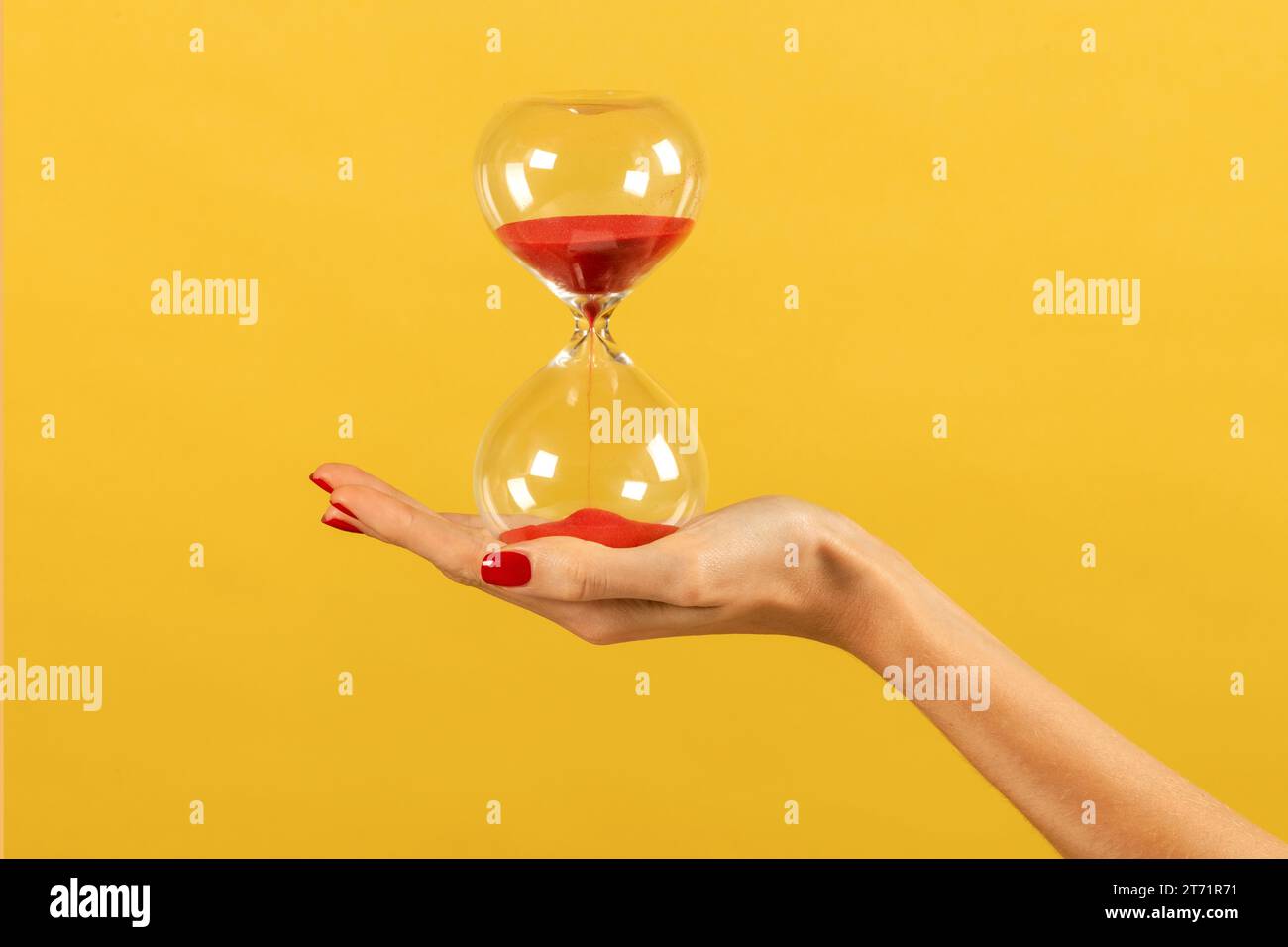 Anonymous young slim female hand with slender fingers and nails polished holding time pass transparent hourglass with red sand in palm against yellow Stock Photo