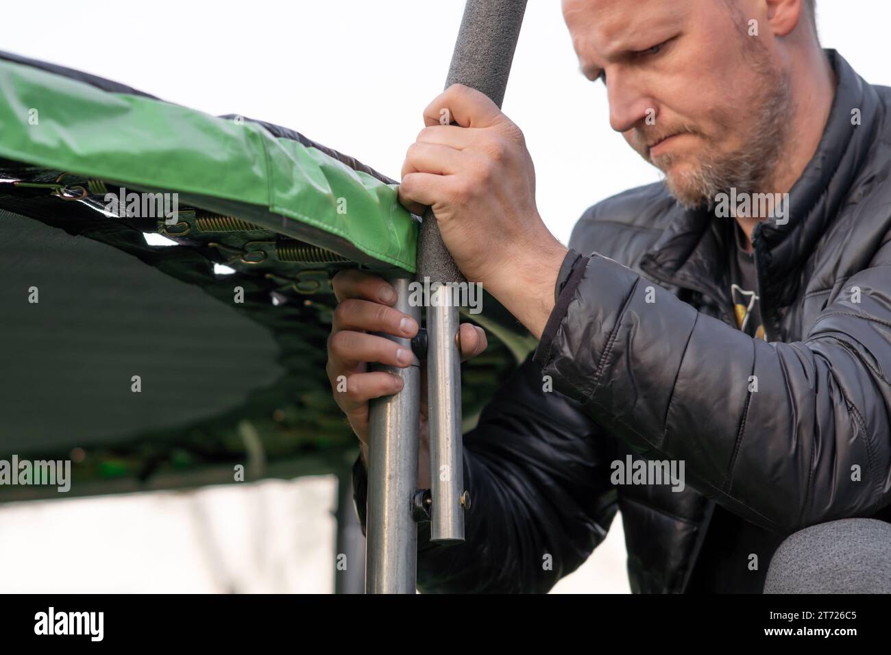 Serious man with beard connecting details of frame for trampoline net with hands and furrowing eyebrows. Mature handyman in black puffy jacket making Stock Photo