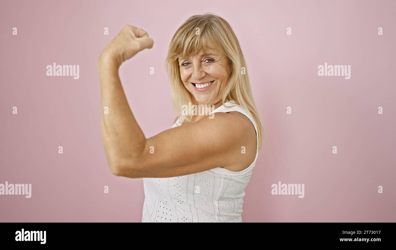 Beautiful middle age blonde woman, beaming with confidence and joy, flexes her strong arm over an isolated pink background. expressing happiness, spor Stock Photo