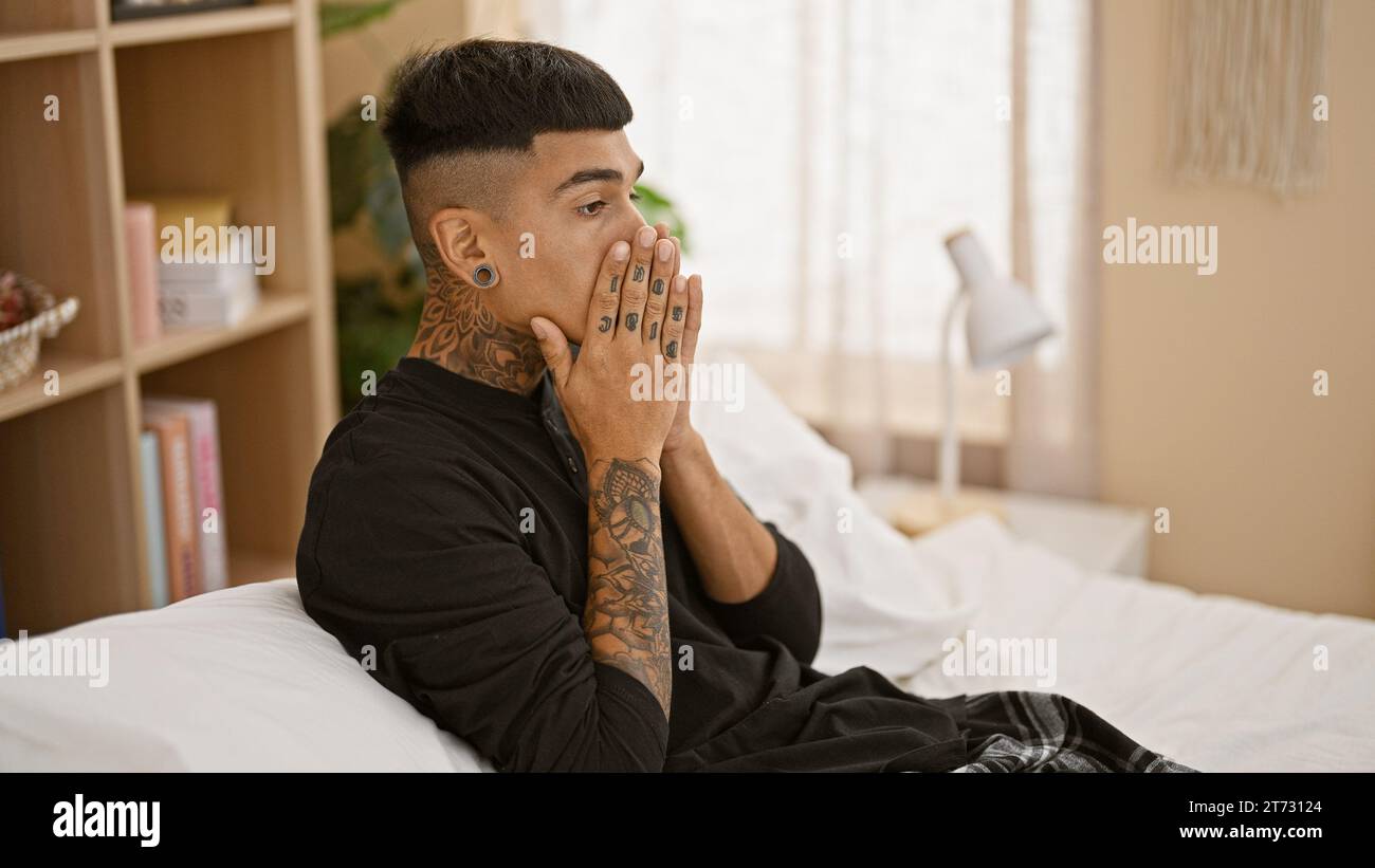 Stressed young latin man sitting on bed, wrestling with anxiety as morning breaks in his bedroom, mirroring the emotional struggle within his tattooed Stock Photo