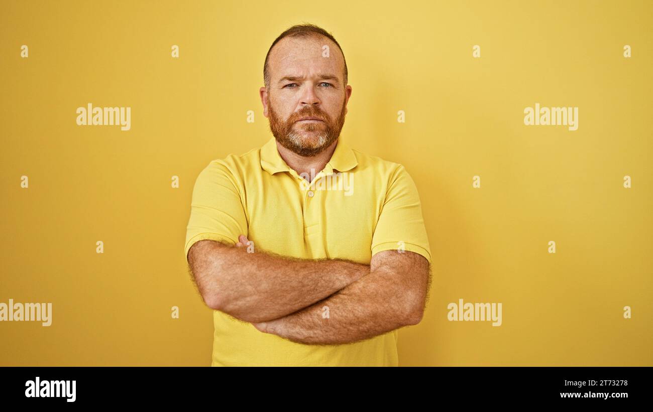 Handsome middle-aged bearded caucasian man in casual getup, standing against a yellow isolated background with arms crossed in a serious expression, c Stock Photo