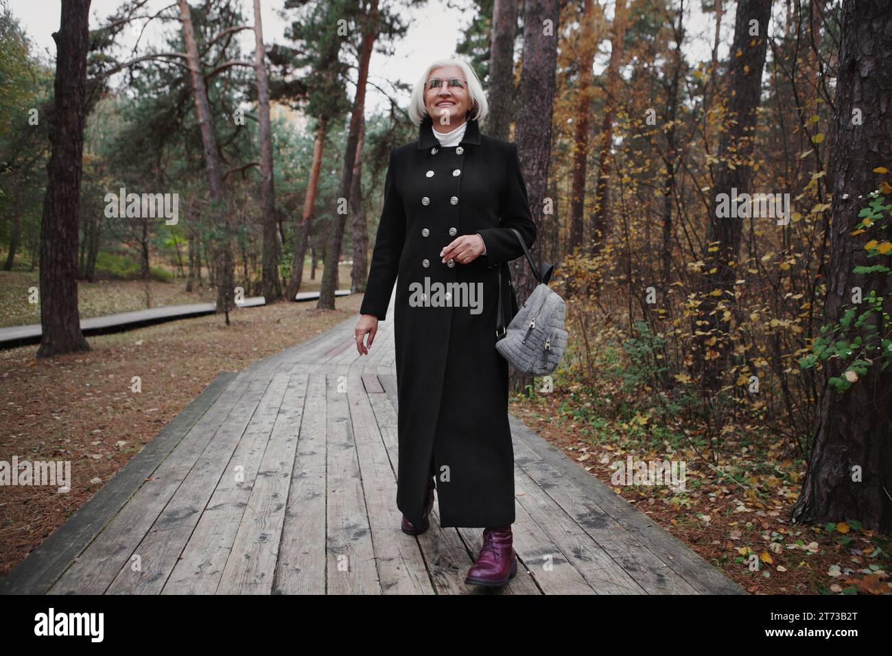 Happy older woman with silver hair and beautiful smile walks through natural park in forest, rejoices and enjoys beauty of nature, genuine and candid Stock Photo