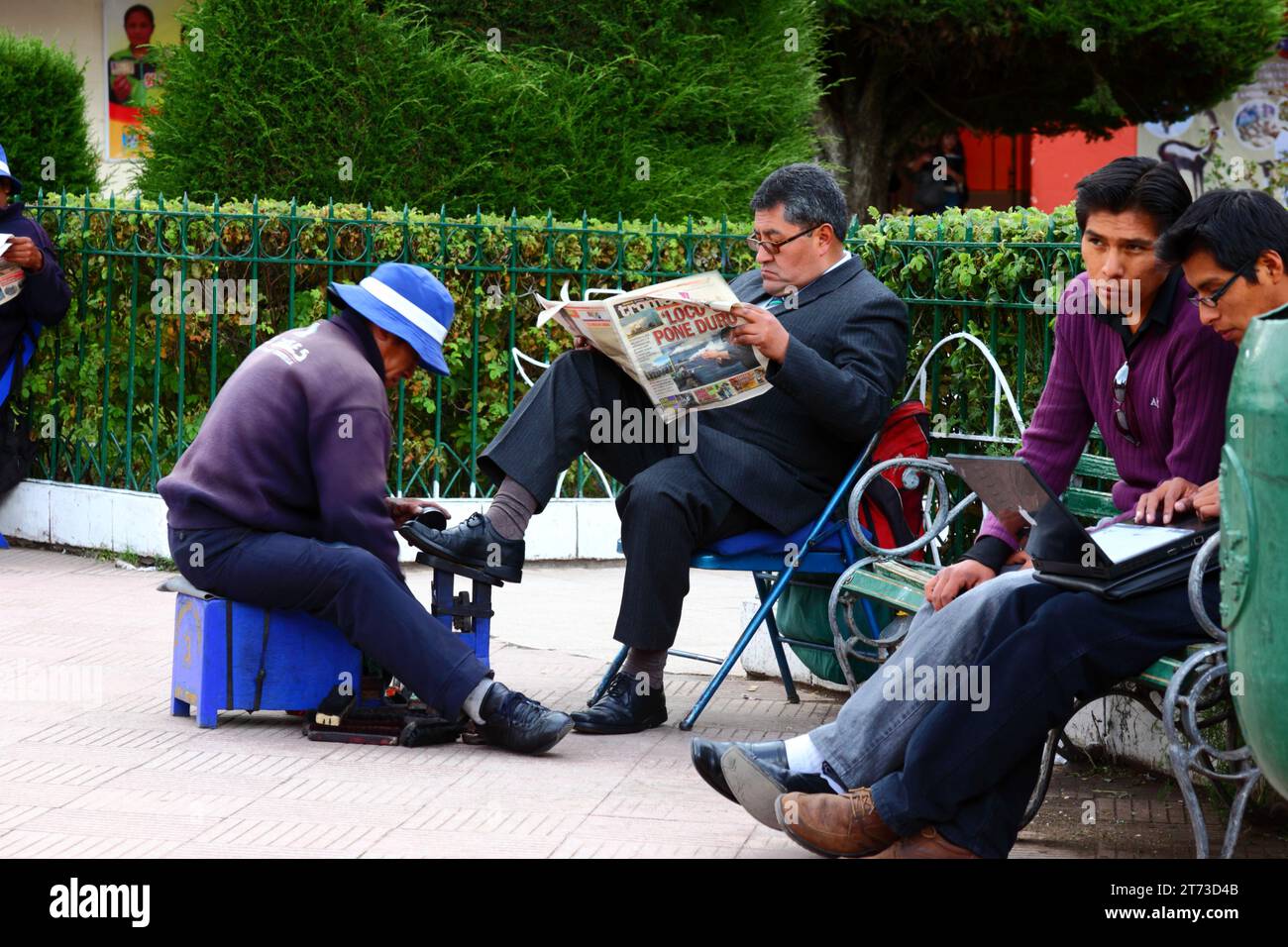 A man wearing a dark suit reads a newspaper while getting his shoes shined in the Plaza de Armas central square, Puno, Peru Stock Photo