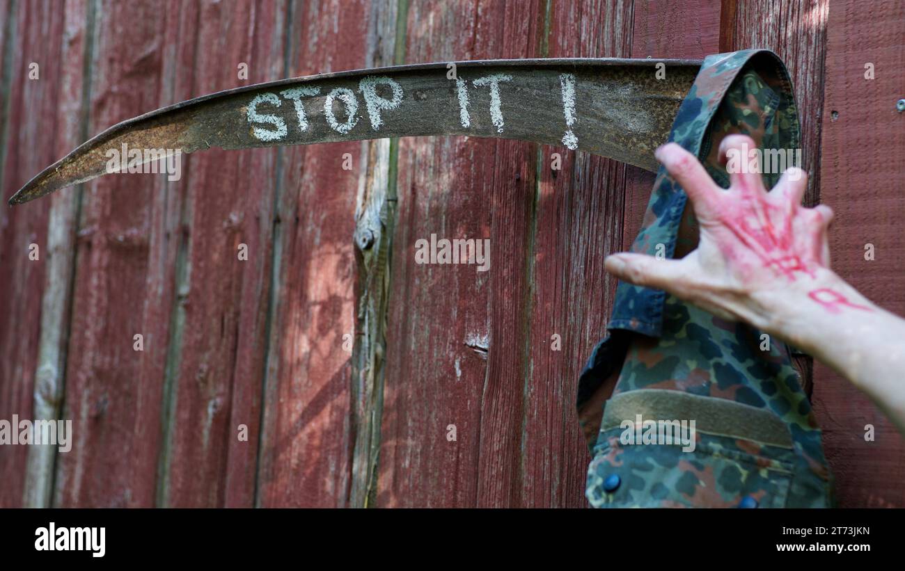 Rusty scythe, STOP IT written on it. A hand with WAR written on it in red grabbing at a military jacket. Stock Photo