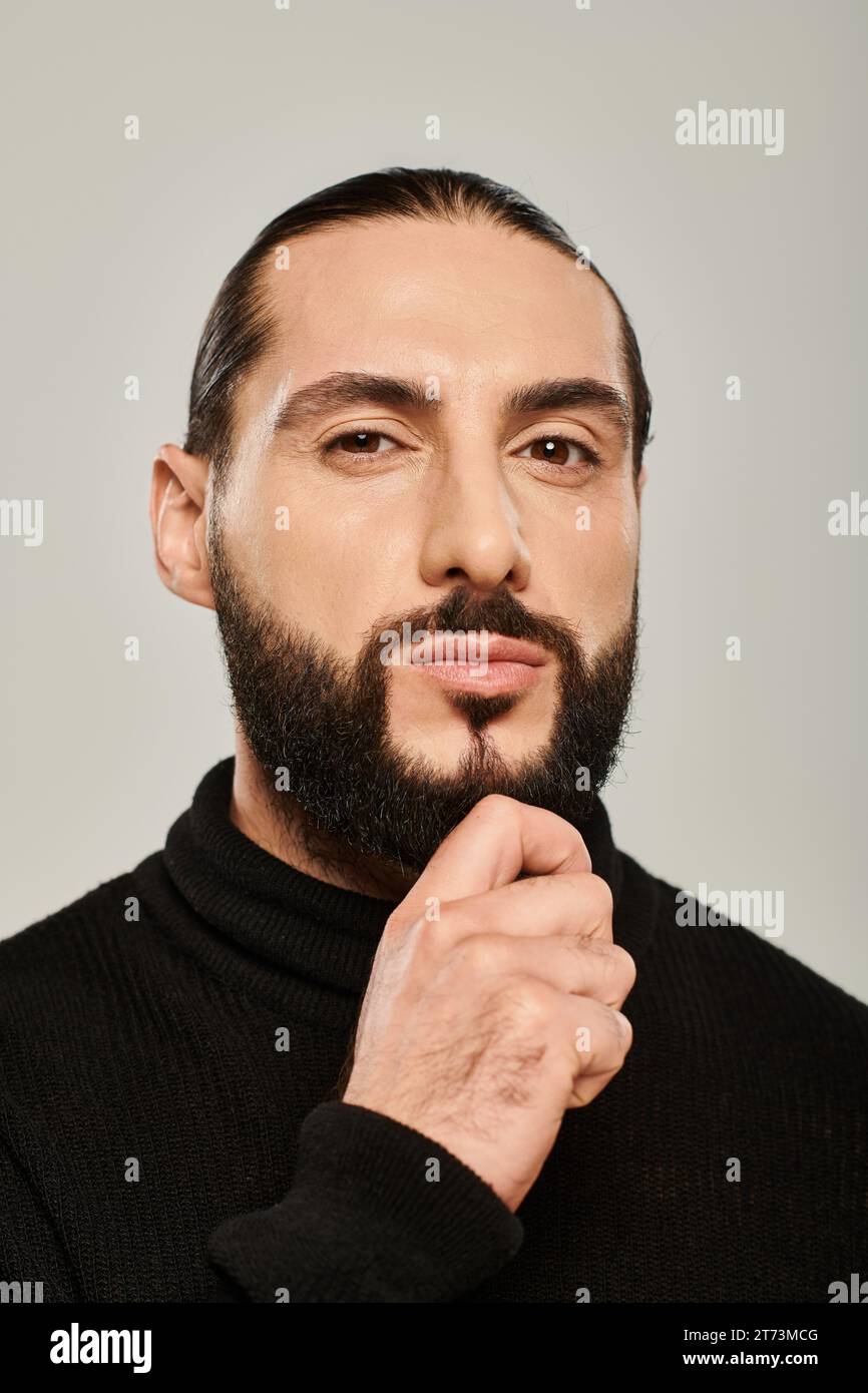 handsome middle eastern man in black turtleneck touching beard on grey background, confidence Stock Photo