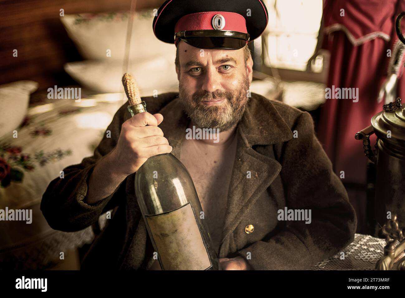 Cossack in a peaked cap and overcoat sits in a hut with a huge bottle of moonshine Stock Photo