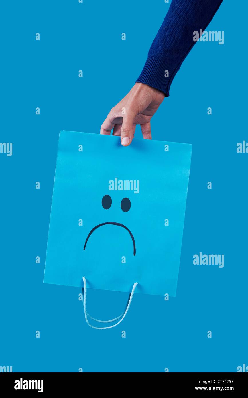 closeup of a man holding an empty blue shopping paper bag, with a sad face painted in it, upside-down on a blue background Stock Photo