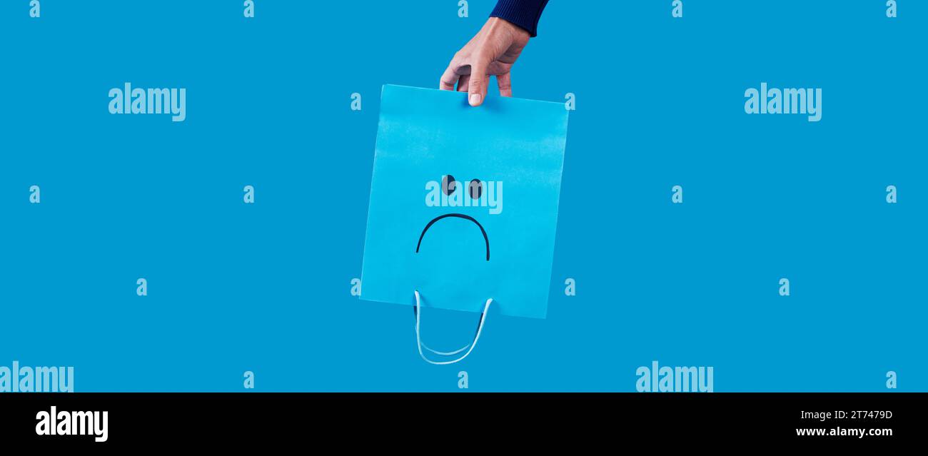 a man holding a blue shopping paper bag, with a sad face painted in it, upside-down on a blue background, in a panoramic format to use as web banner Stock Photo