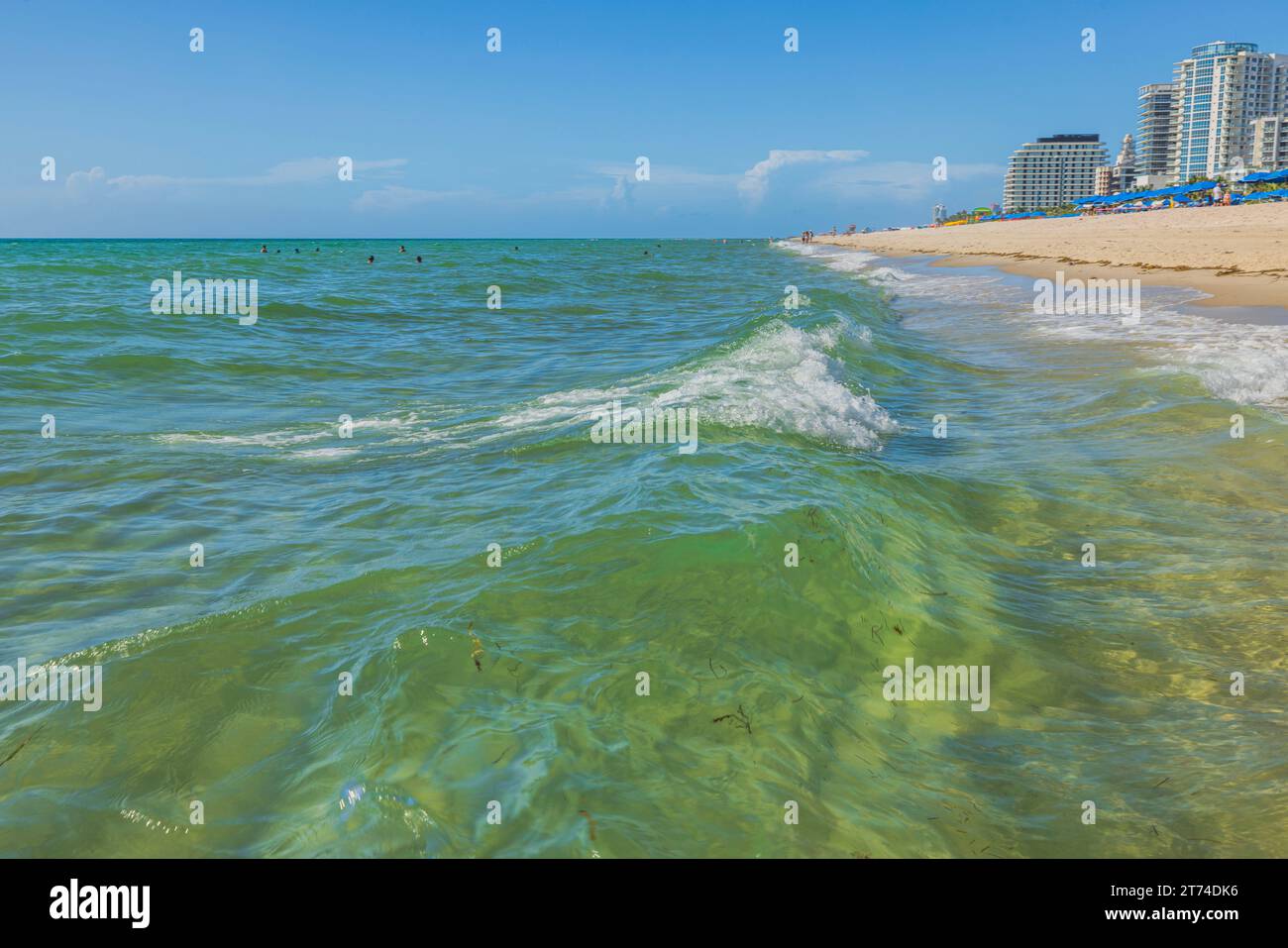 View of shoreline with architectural buildings under blue sky on horizon accompanied by gentle waves rolling onto sandy beach. Miami Beach, Florida, Stock Photo