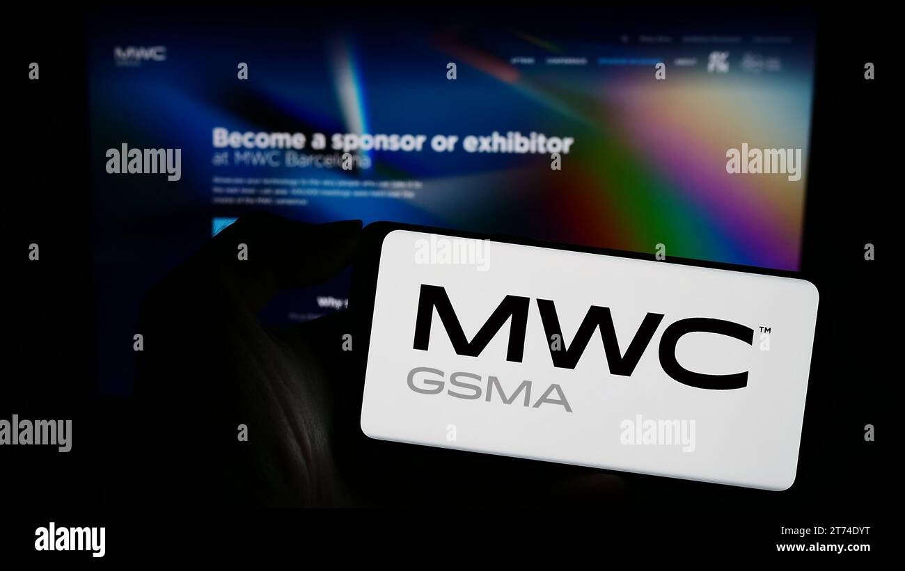 Person holding cellphone with logo of annual mobile communications trade show MWC Barcelona in front of business webpage. Focus on phone display. Stock Photo