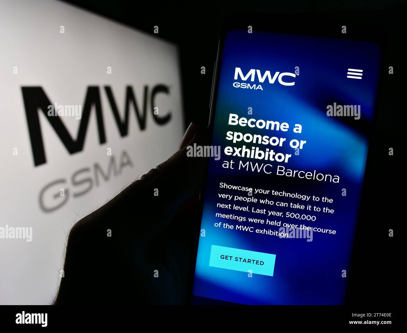 Person holding cellphone with website of annual mobile communications trade show MWC Barcelona in front of logo. Focus on center of phone display. Stock Photo