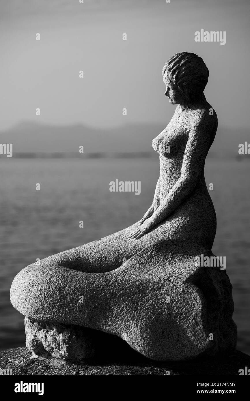 A white marble statue of a figure is perched on a rock facing a serene lake, the sun reflecting off the placid surface of the water Stock Photo