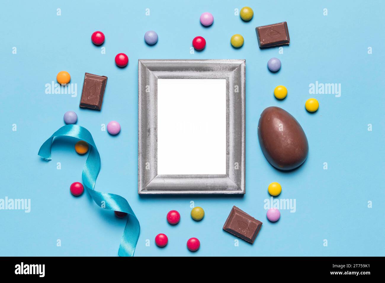 Empty blank white silver frame surrounded with easter egg gem candies chocolate pieces blue backdrop Stock Photo