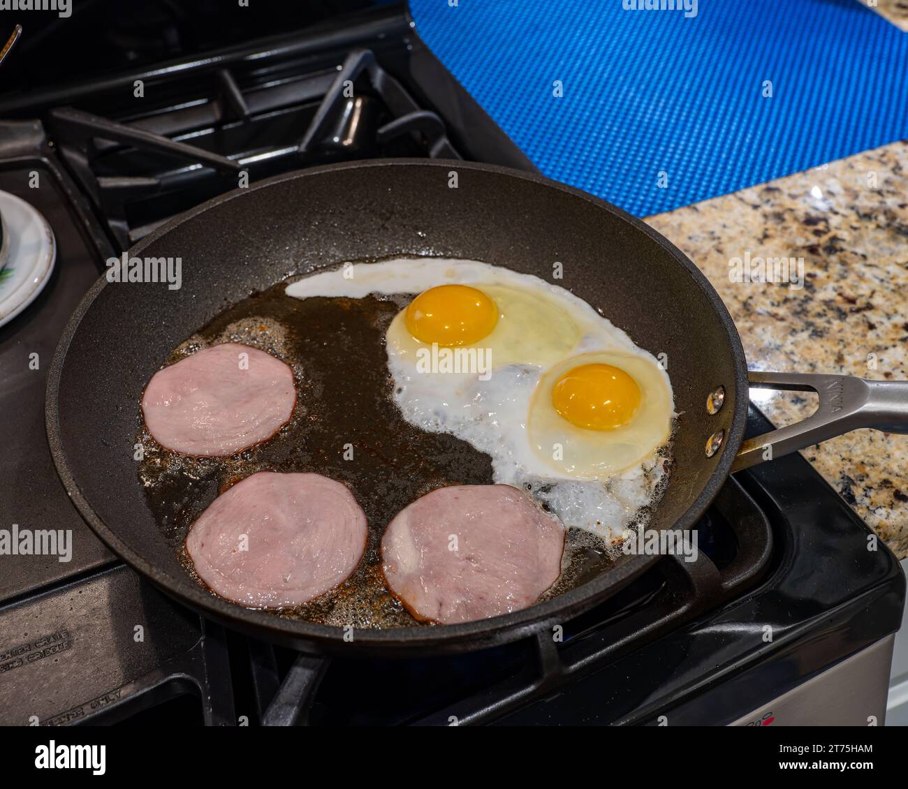 Fried or frying eggs with Canadian bacon in a skillet in home kitchen on gas stove top, a typical American breakfast. Stock Photo