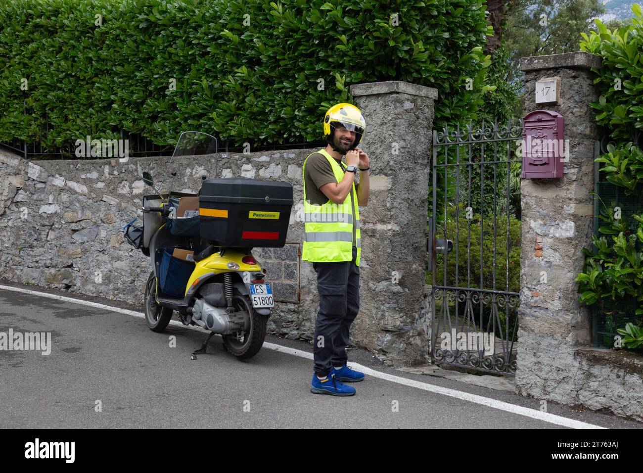 A Poste Italiane mail carrier fastens his helmet after parking his motorbike along a wall in Bellagio, Lombardy, Italy. Stock Photo