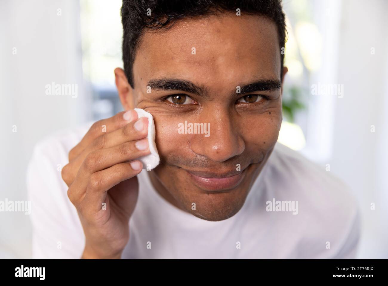 Happy biracial man cleansing face with cotton pad in bathroom at home Stock Photo