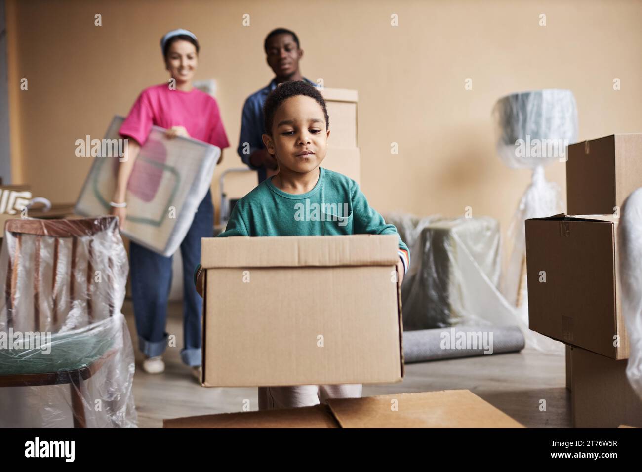 Cute little boy carrying packed cardboard box and putting it on top of another one while helping his parents with relocation to new apartment Stock Photo