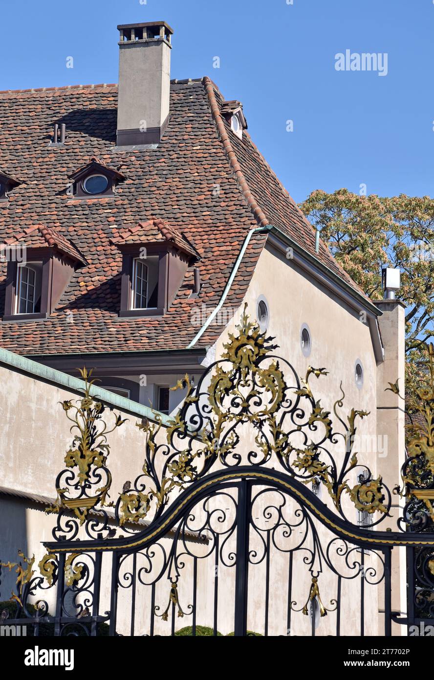 Elegant wrought iron gates topped with elaborate gilded work, and beyond a tiled gabled roof with several gabled windows Stock Photo
