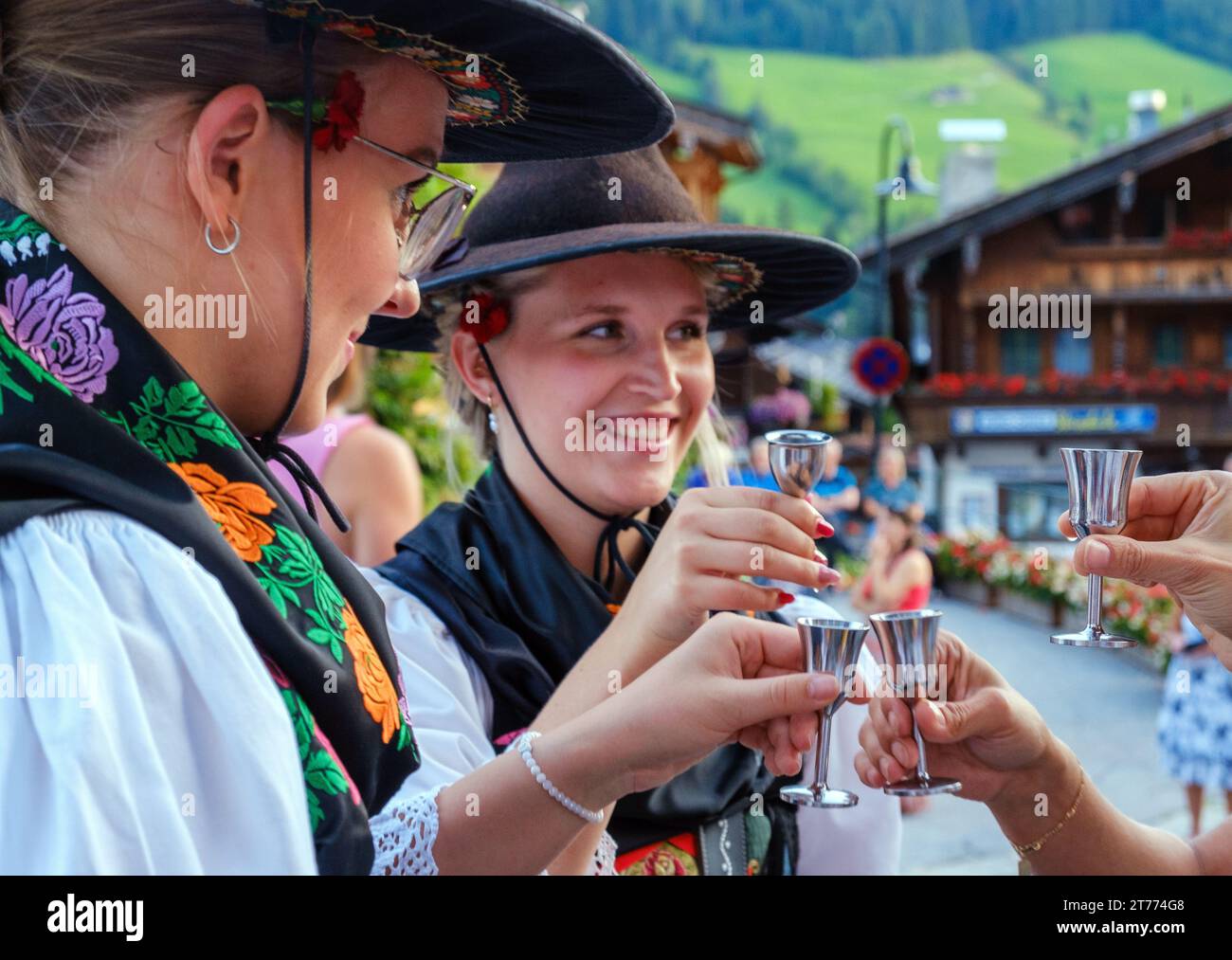 Two females in traditional Tyrolean dress, hold up silver Schnaps goblets, toasting two tourists in Alpbach, Austria. Stock Photo