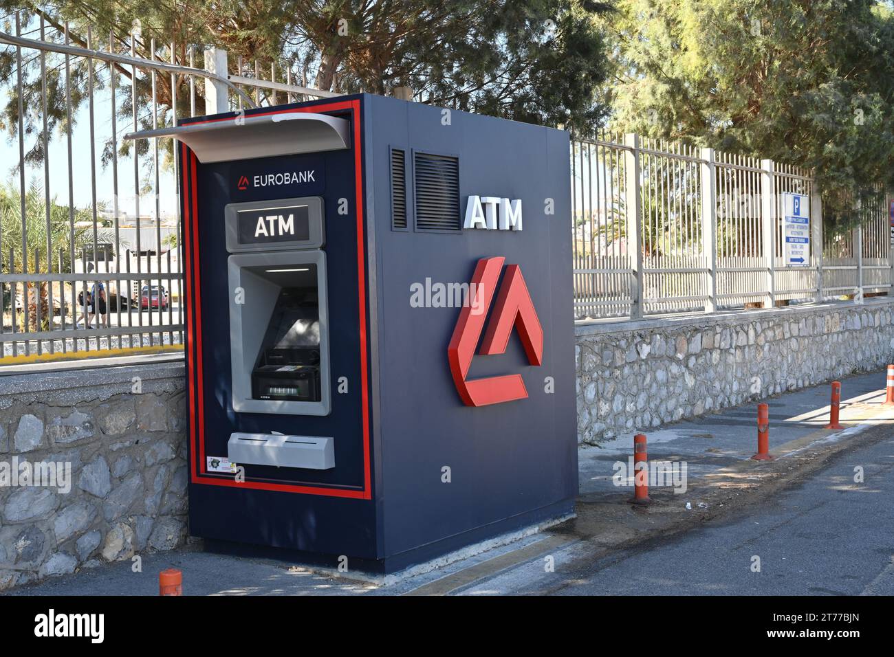 Blue ATM machine of Eurobank is standing alone in the area of passenger seaport. Behind is metal fence and small stone wall Stock Photo