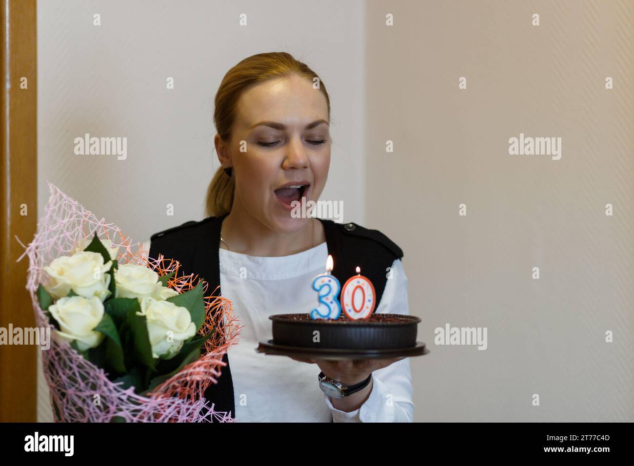 A young happy woman with a bouquet of flowers in her hands blows out the candles on the cake, celebrates her thirtieth birthday. Stock Photo