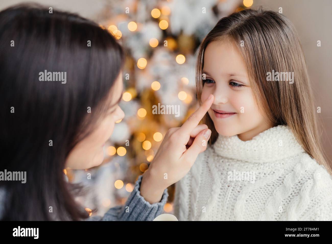 A tender moment as a mother affectionately touches her child's nose, with a softly lit Christmas tree behind Stock Photo