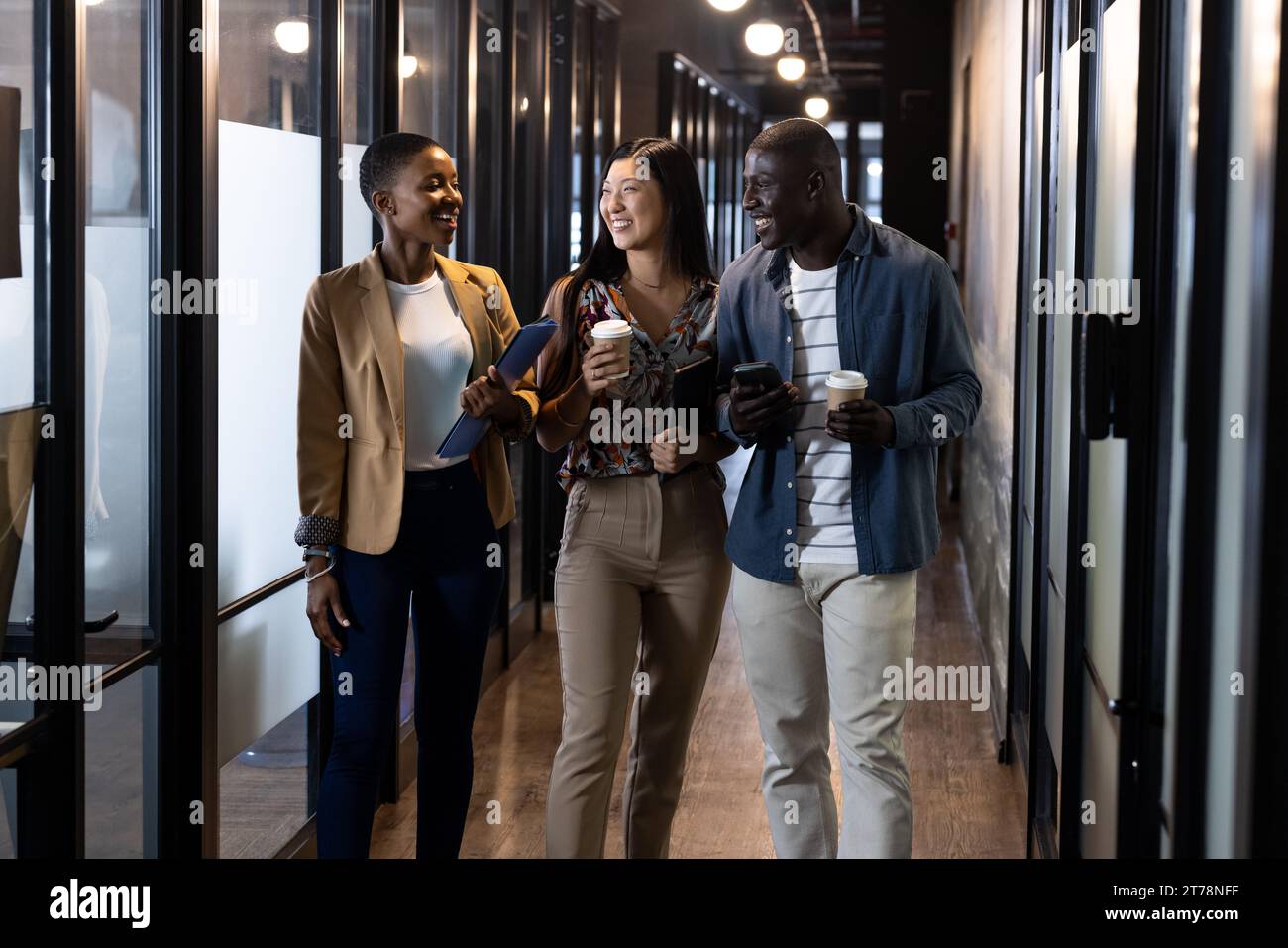 Happy diverse male and female colleagues with coffees, tablet and smartphone talking in corridor Stock Photo