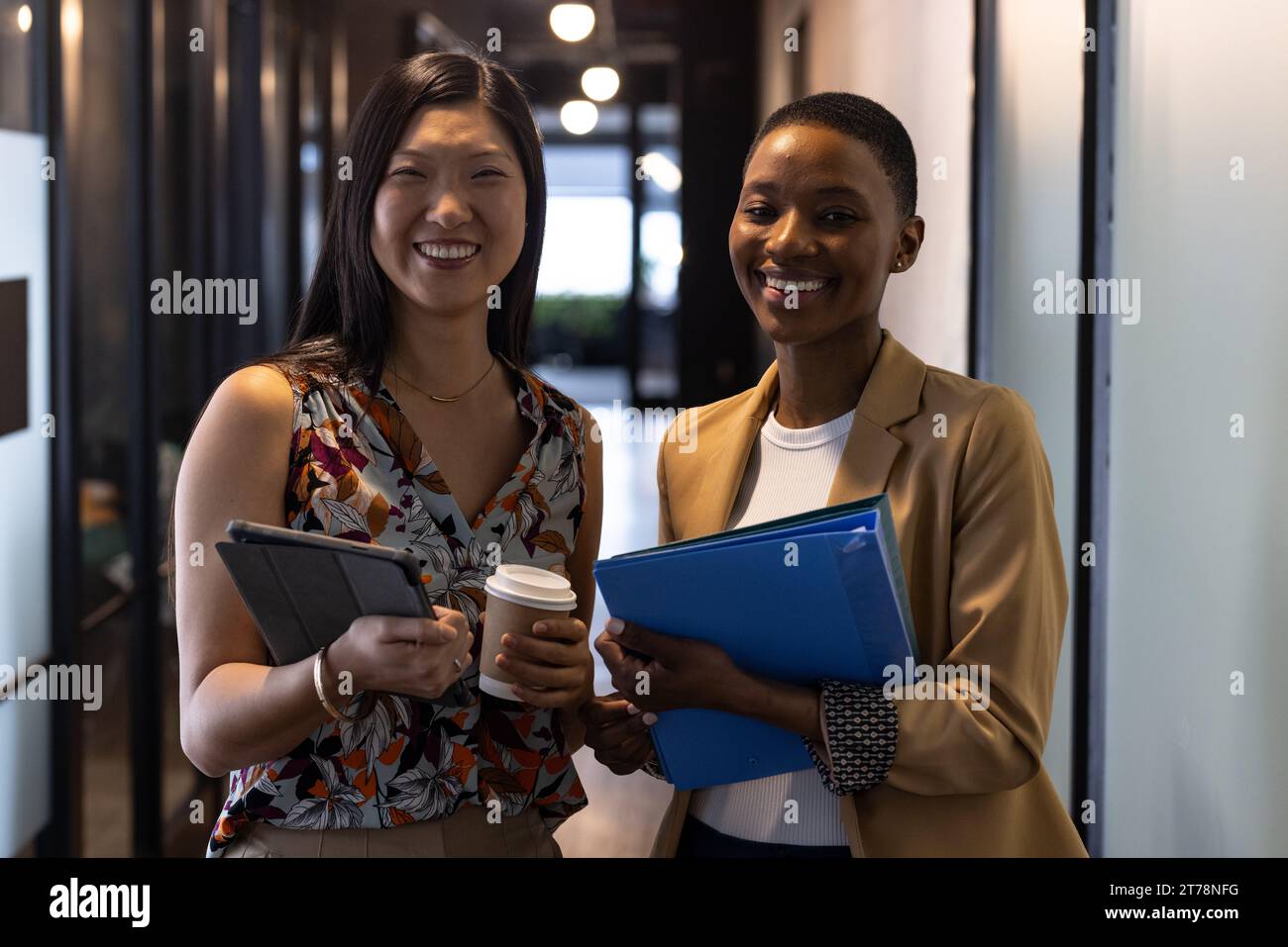 Portrait of happy diverse female colleagues with coffees, tablet and paperwork smiling in corridor Stock Photo