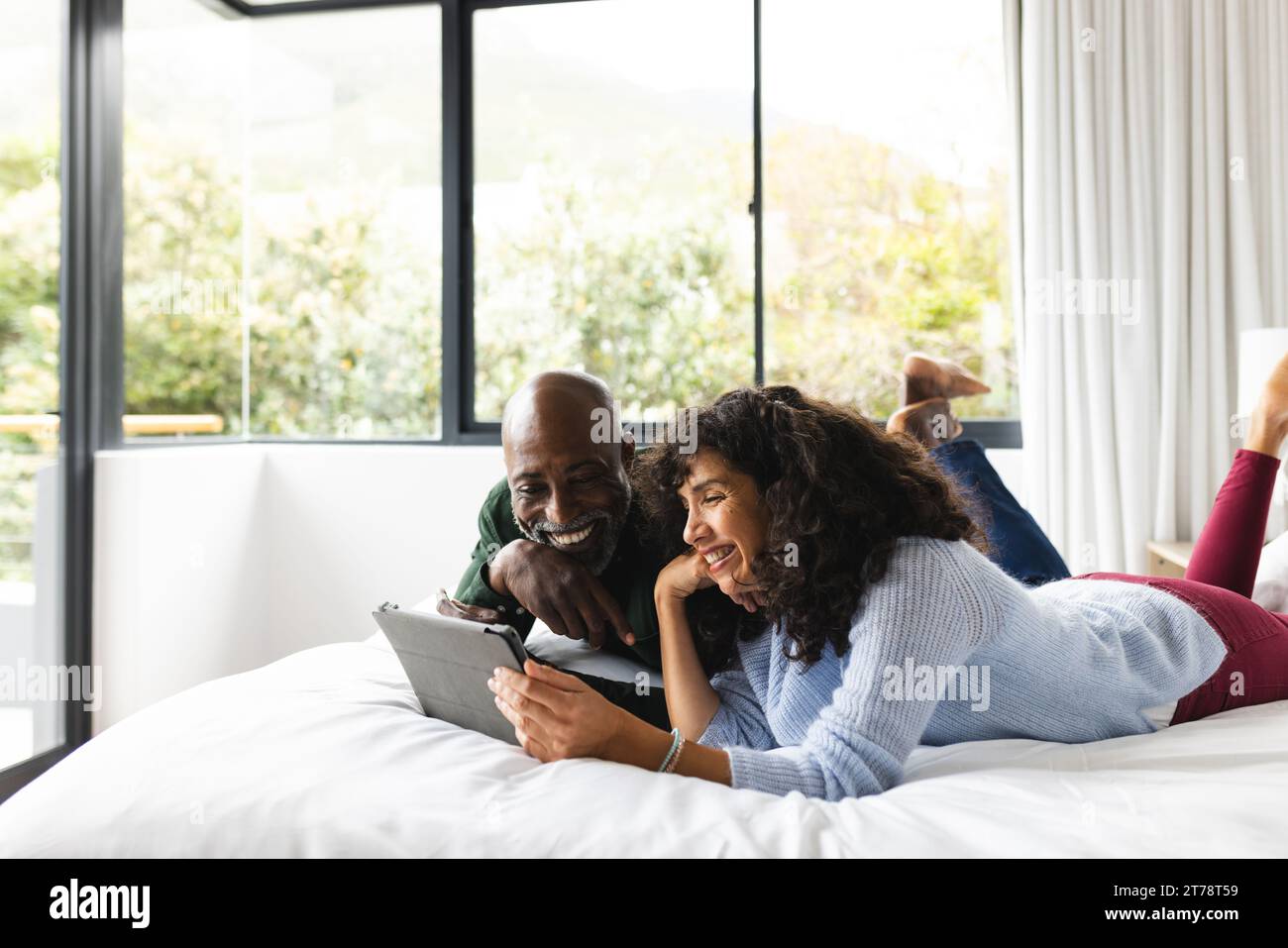 Happy diverse mature couple lying on bed using tablet in sunny bedroom, copy space Stock Photo