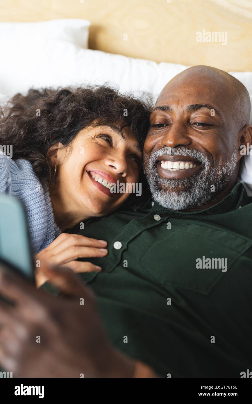 Happy diverse mature couple lying on bed embracing using tablet in sunny bedroom Stock Photo