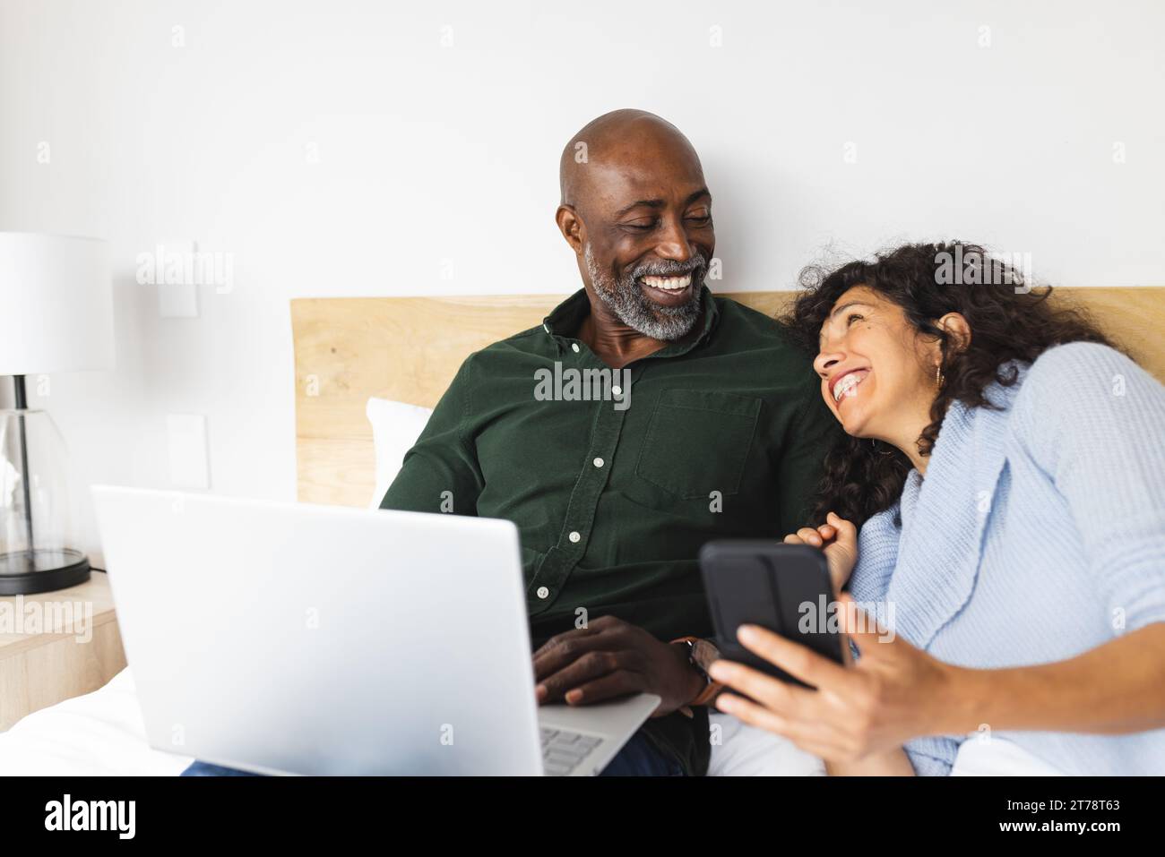 Happy diverse mature couple sitting on bed using laptop and smartphone in bedroom Stock Photo