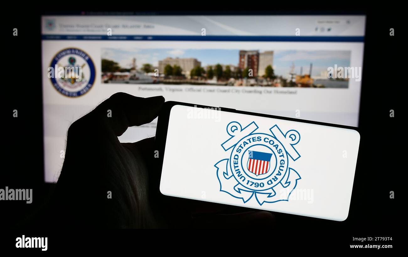 Person holding cellphone with seal of United States Coast Guard (USCG) in front of webpage. Focus on phone display. Stock Photo