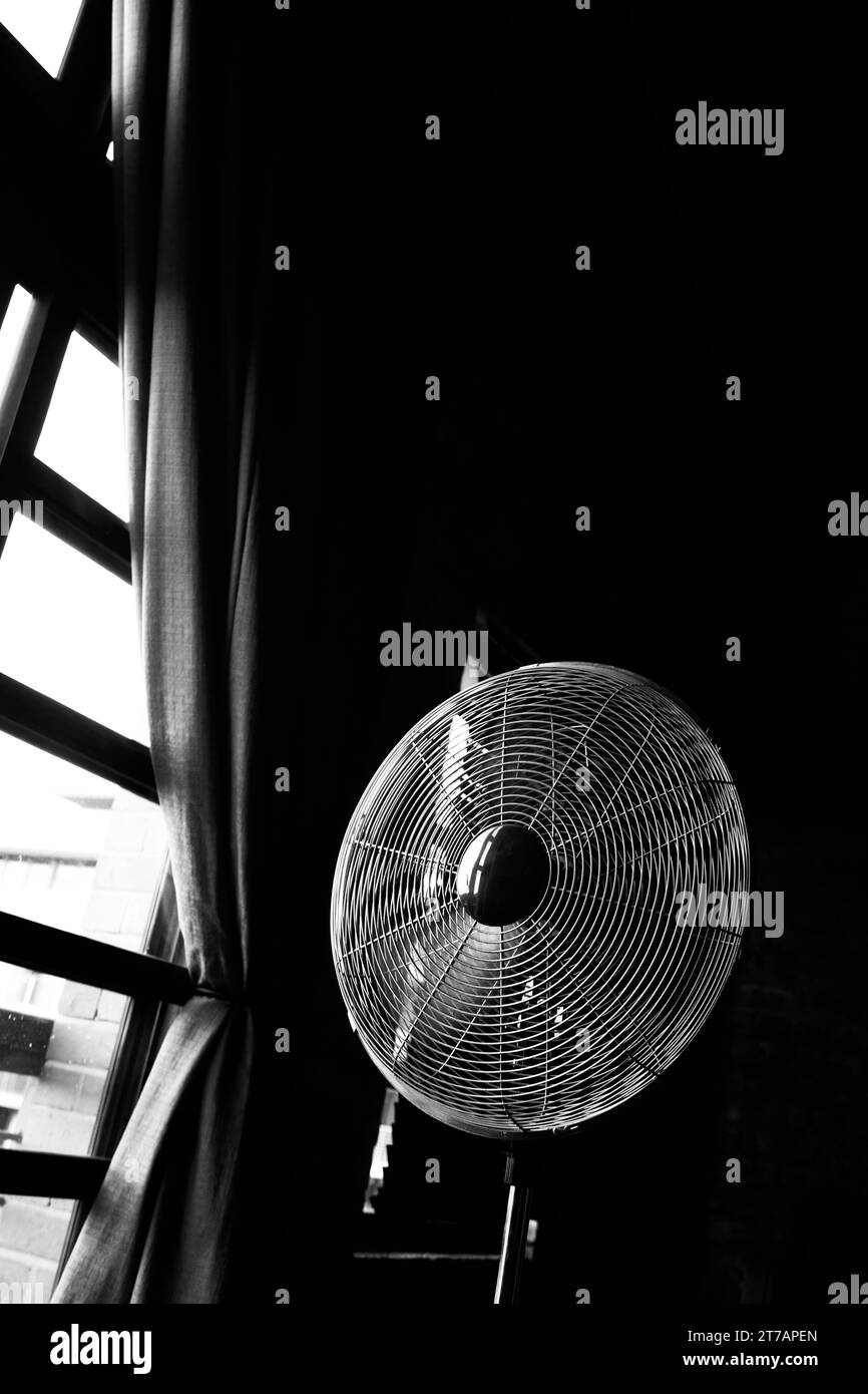Black and white photo of a vintage floor fan standing by a tall window