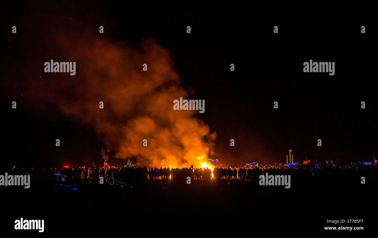 A group of people of various ages and ethnicities enjoying a nighttime outdoor gathering around a large bonfire in a picturesque outdoor setting Stock Photo