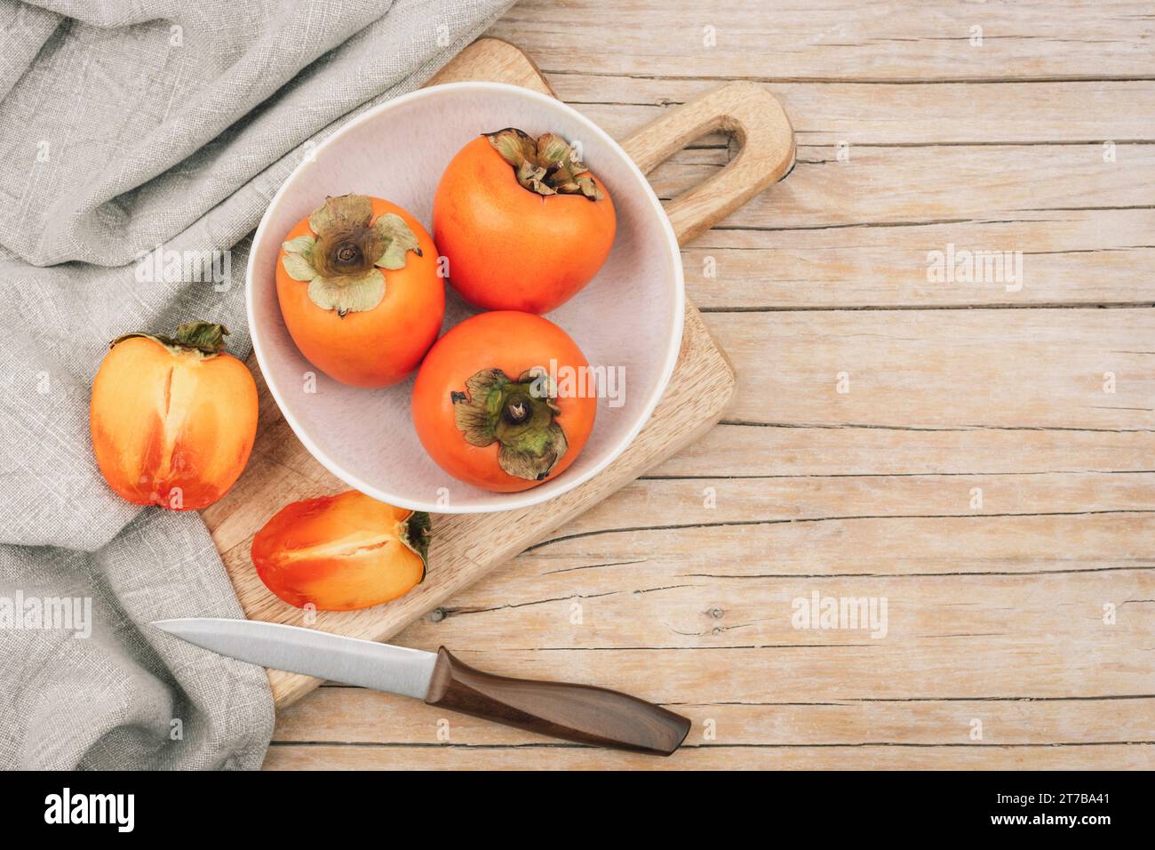 Fresh ripe persimmons in white bowl on cutting board on wooden table. Healthy vegan food concept. Top view, flat lay, mockup. Stock Photo