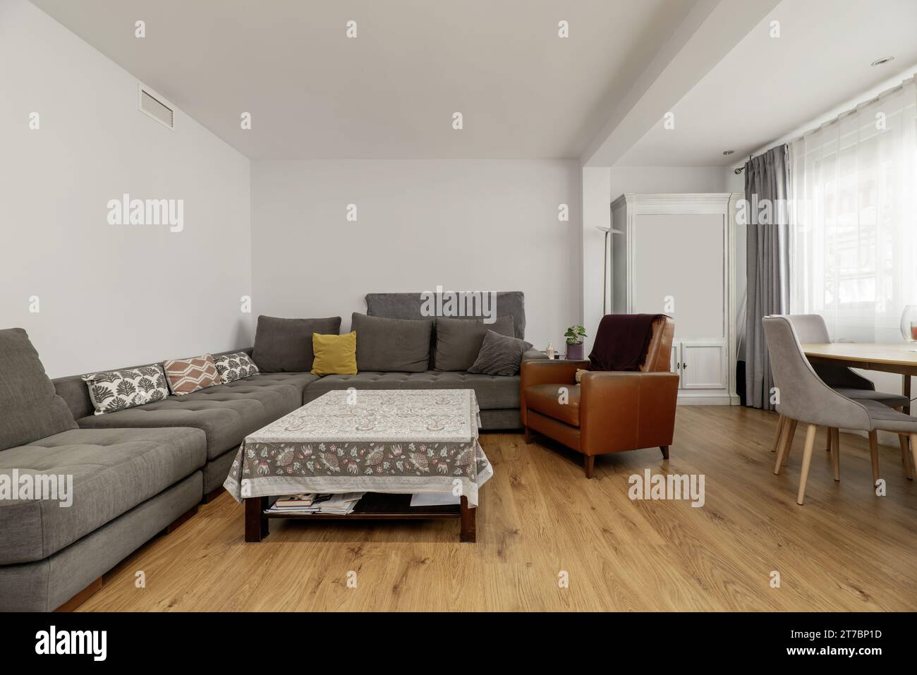 A spacious living room with a corner sofa upholstered in gray fabric, a square coffee table with a tablecloth, Stock Photo