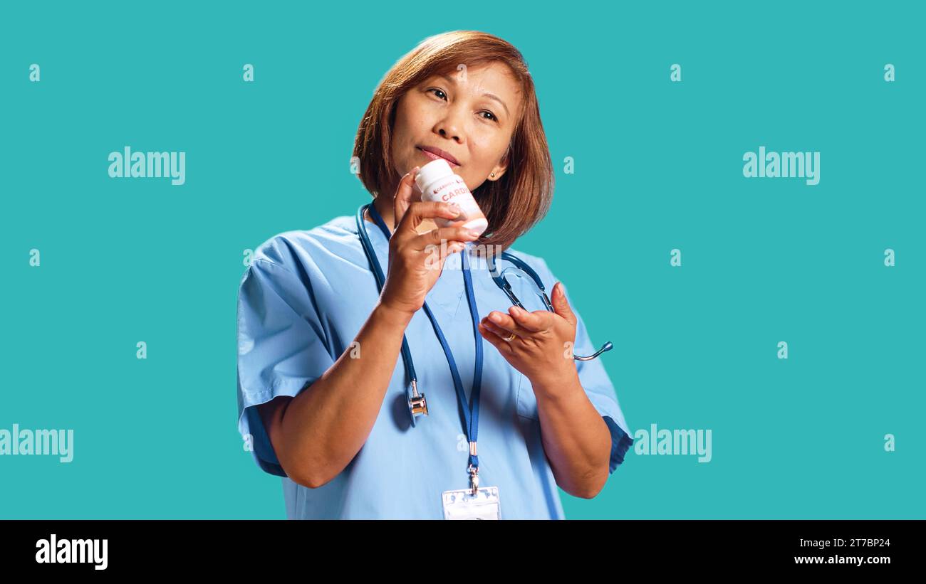 Cheerful skilled nurse holding cardiovascular bottle pill, offering medical guidance, close up. Hospital worker prescribing heart antibiotic to patient, isolated over blue studio background Stock Photo