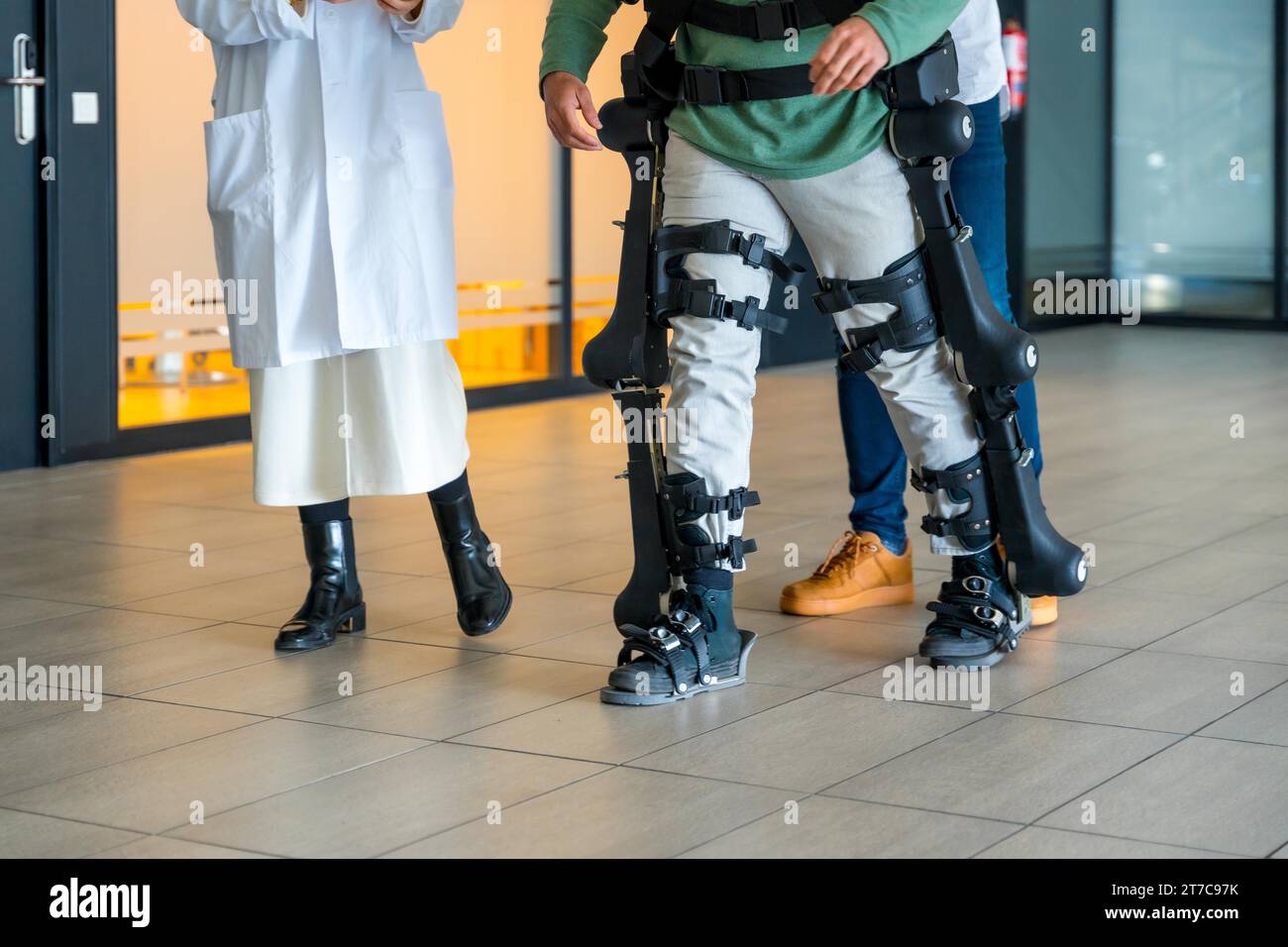 Mechanical exoskeleton, female doctor with unrecognizable engineer walking with disabled person with robotic skeleton in rehabilitation Stock Photo