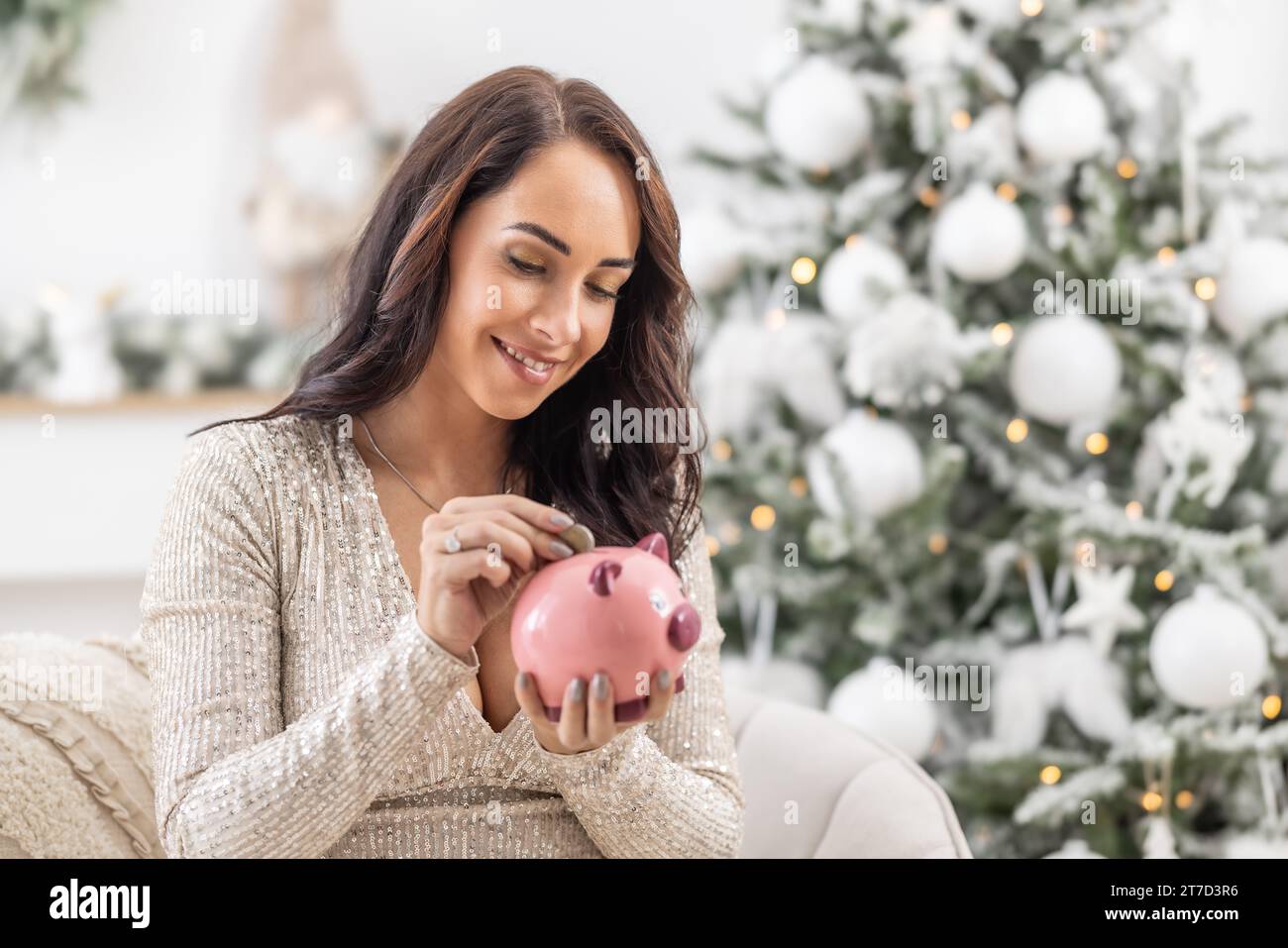 Smiling woman puts a coin into a pink piggybank sitting next to a Christmas tree. Stock Photo