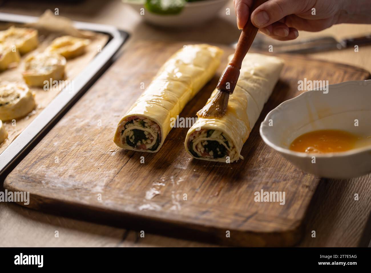 The cook brushes an egg onto a puff pastry roll. Recipe procedure 7 of
