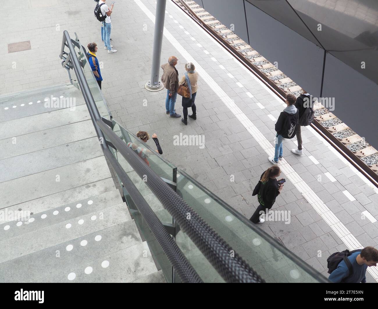 View from the staircase down to railway track with people waiting, older couple, girl, boy, headphones, handrail, safety strip, Utrecht Centraal Stock Photo