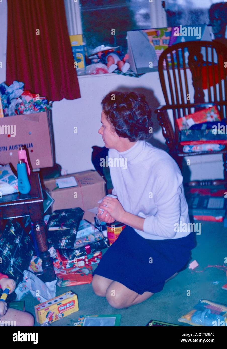 A 1960s mother clearing up discarded Christmas wrapping paper. Some presents including toys are open; others are still wrapped. A box for the rubbish in the background. Northamptonshire 1968, Christmas Day Stock Photo