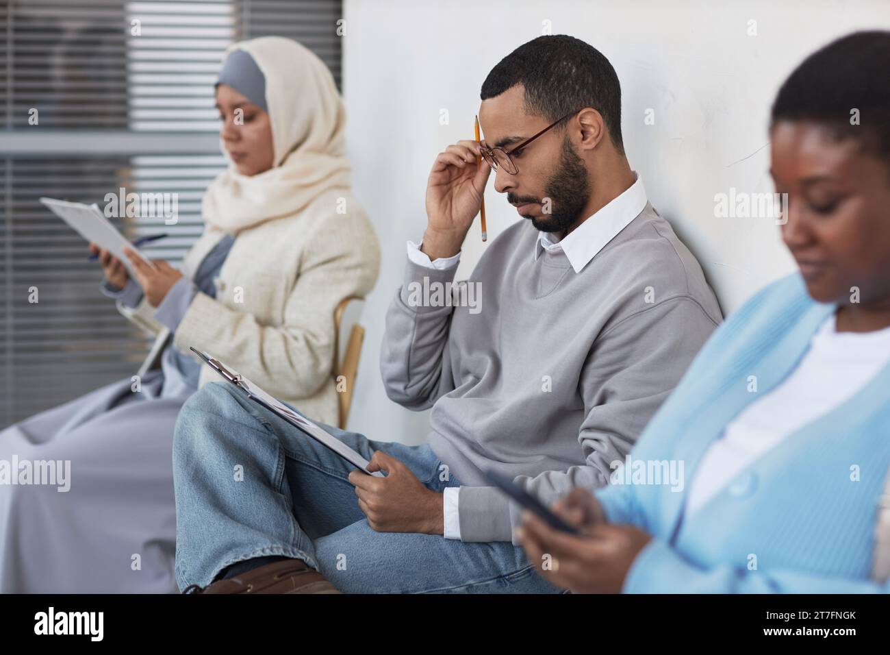 Focus on young serious multiethnic man looking through questions of visa application form and thinking of answers while sitting in queue Stock Photo