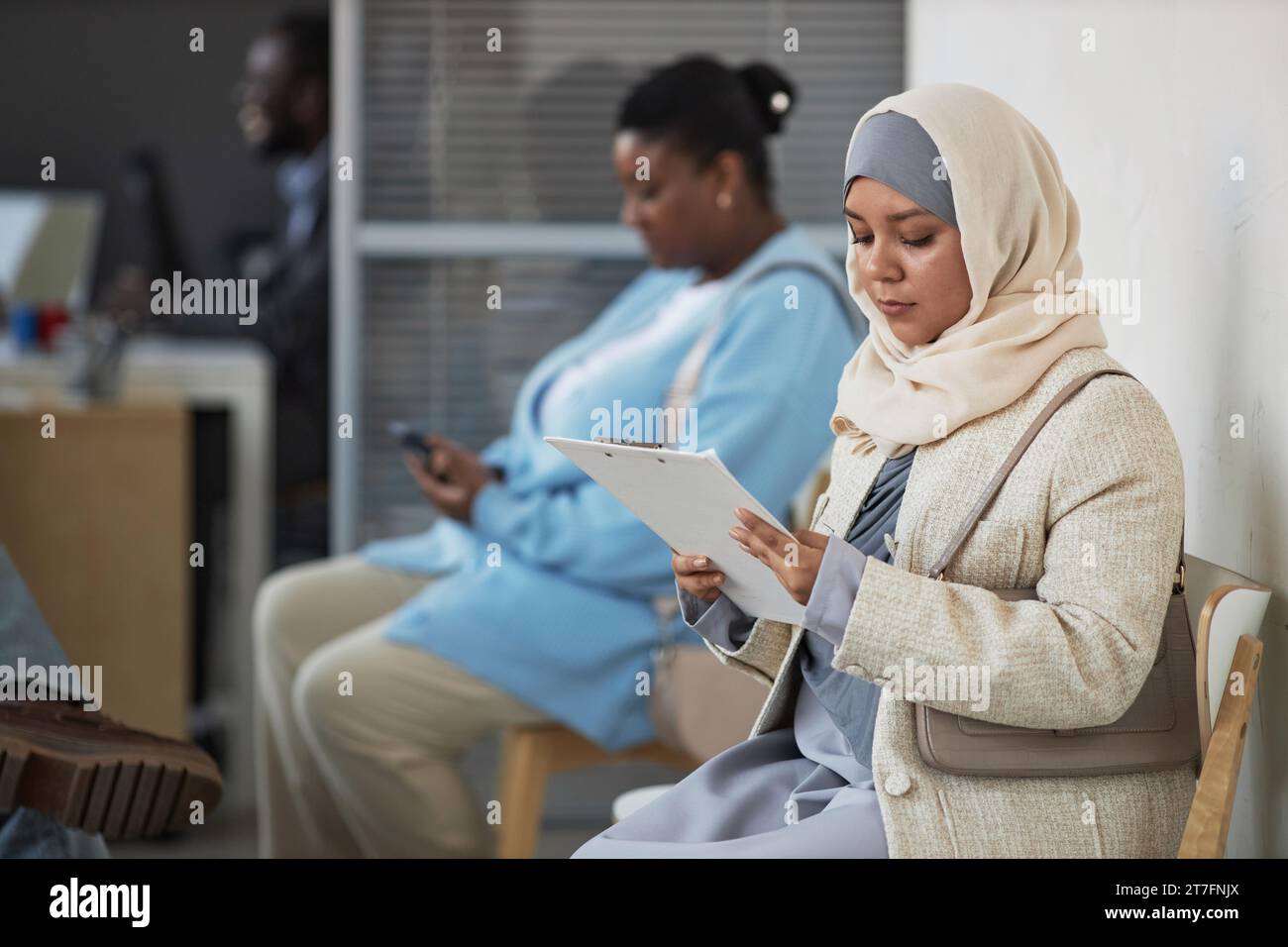Young Muslim woman in hijab reading application form while sitting in queue to manager office in visa request center Stock Photo