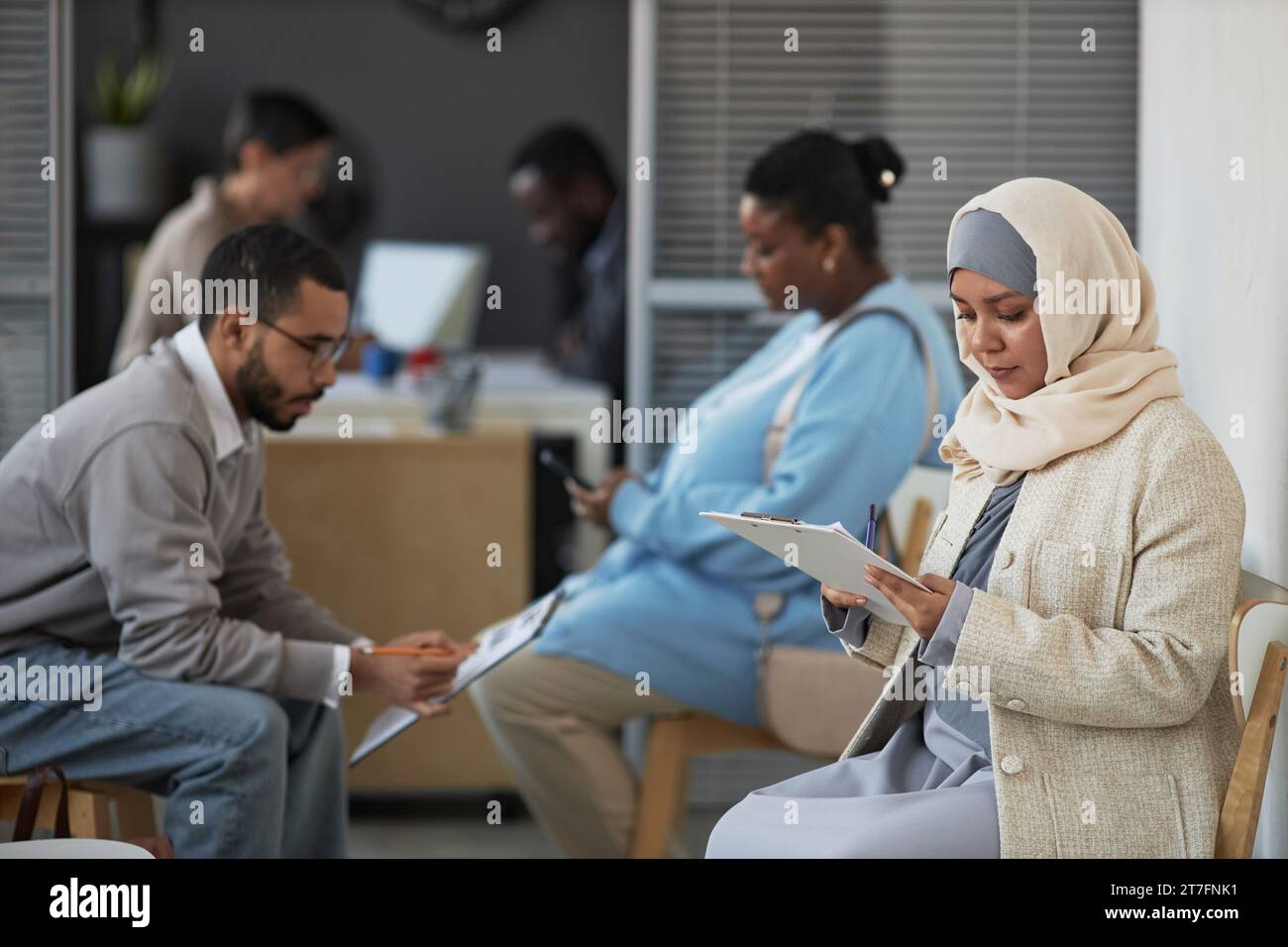 Young serious female applicant in hijab reading questions of visa application form while filling it in against other people Stock Photo