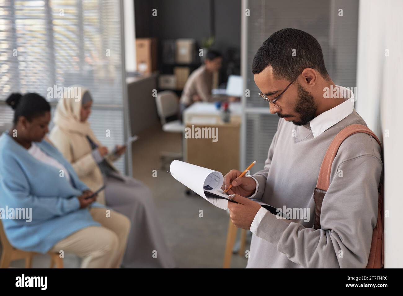 Young male applicant filling in visa application form in front of camera while standing by wall and waiting for his turn to manager office Stock Photo