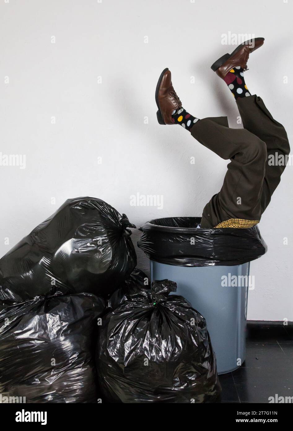 Businessman in Suit and Polk-A-Dot Socks Stuck Upside Down in Trash Can Next to Garbage Bag Pile. Concept of Over a Barrel. Out of Fashion. Stock Photo