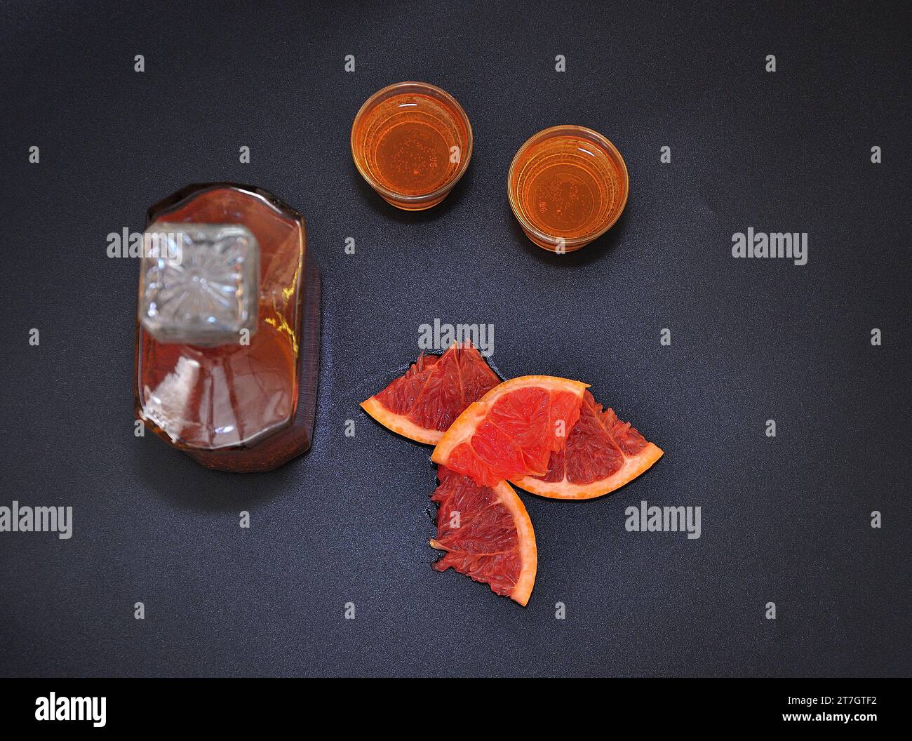 Grapefruit liqueur on a black background, pieces of ripe citrus, a glass decanter and two glasses of strong alcohol on a black background. Top view, f Stock Photo